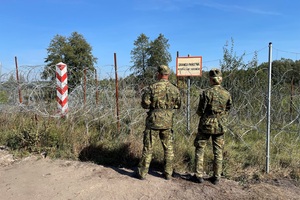 Patrol Straży Granicznej W tle rzeka i bariera fizyczna, przed nią tabliczka granica państwa oraz dwóch funkcjonariuszy Straży Granicznej, stoją plecami.