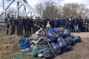 Akcja sprzątania rzeki Biebrzy Duża grupa osób stojąca na zewnątrz, pozuje do wspólnego zdjęcia. Przed nimi leży duża sterta zebranych śmieci w workach oraz różnych odpadów. W tle znajdują się drzewa i metalowy most.