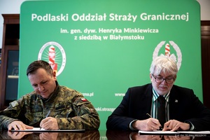 Współpraca Podlaskiego Oddziału Straży Granicznej i Muzeum Wojska Polskiego w Białymstoku Zdjęcie przedstawia dwóch mężczyzn siedzących przy stole. Obaj są uchwyceni w momencie podpisywania dokumentów.